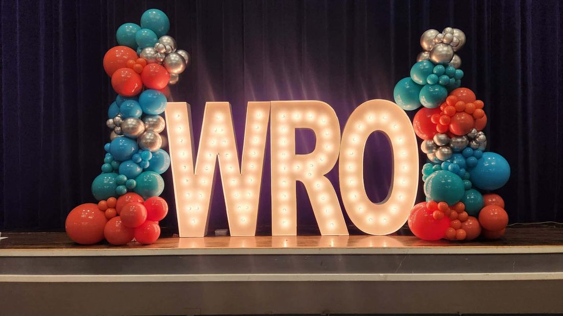 Illuminated marquee letters flanked by teal and coral balloon garlands on a stage