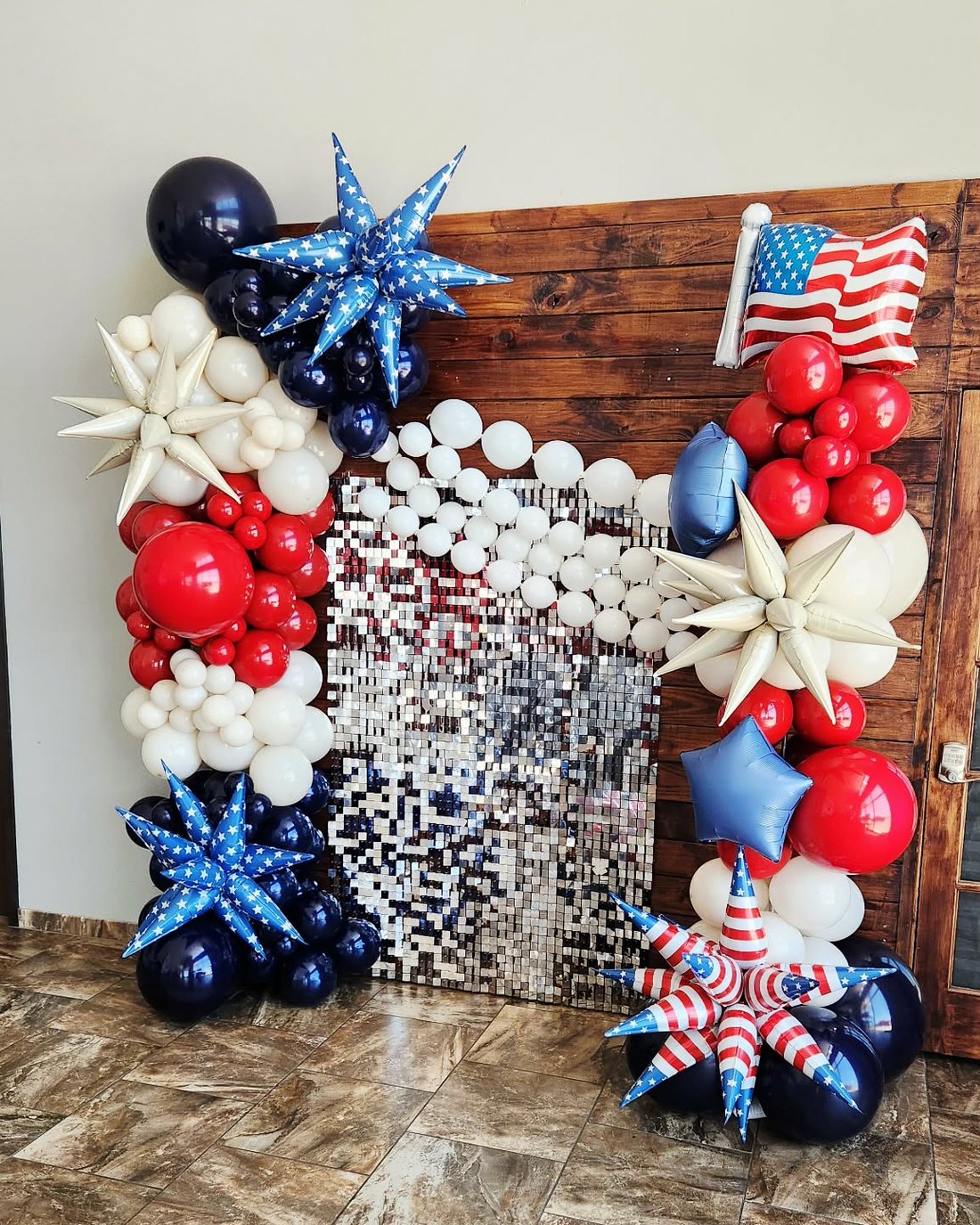 Fourth of July balloon installation in red, white and navy with star bursts and a flag balloon, framing a silver sequin wall on rustic wood