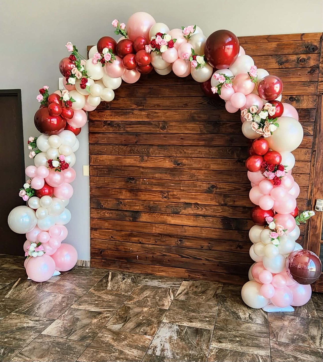 Blush, ivory and burgundy balloon arch accented with roses, set against a rustic wood backdrop